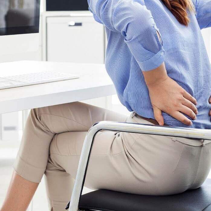 의자에 앉아있는 하늘색 셔츠를 입은 여성이 통증이 있는 듯이 허리를 잡고 있는 모습