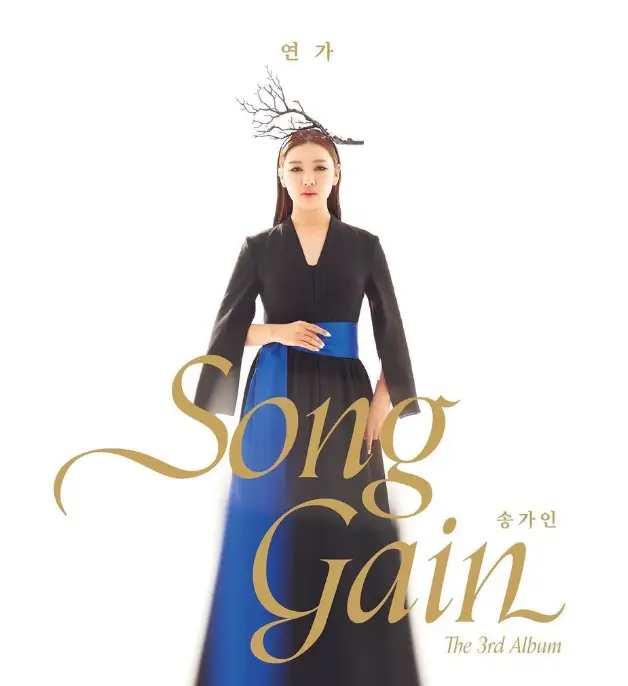 송가인- 흰배경 검은 긴팔 드레스를 입고 무표정하게 서있는 송가인 앞 황금글씨 songgain