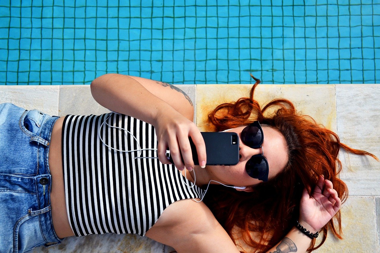 수영장에 누워서 이어폰으로 음악을 듣고있는 줄무늬 민소매를 입고 썬글라스를 쓰고 있는 여성