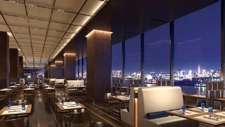 창밖으로 불빛을 내고 있는 빌딩이 보이고 내부에는 테이블과 의자가 많이 세팅되어 있는 식당