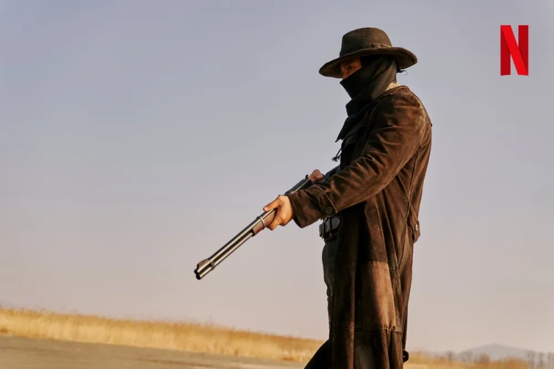 갈대를 배경으로 카우보이 모자에 긴 갈색 롱코트와 장총을 들고 있는 넷플릭스 드라마 도적 칼의 소리의 이윤 캐릭터