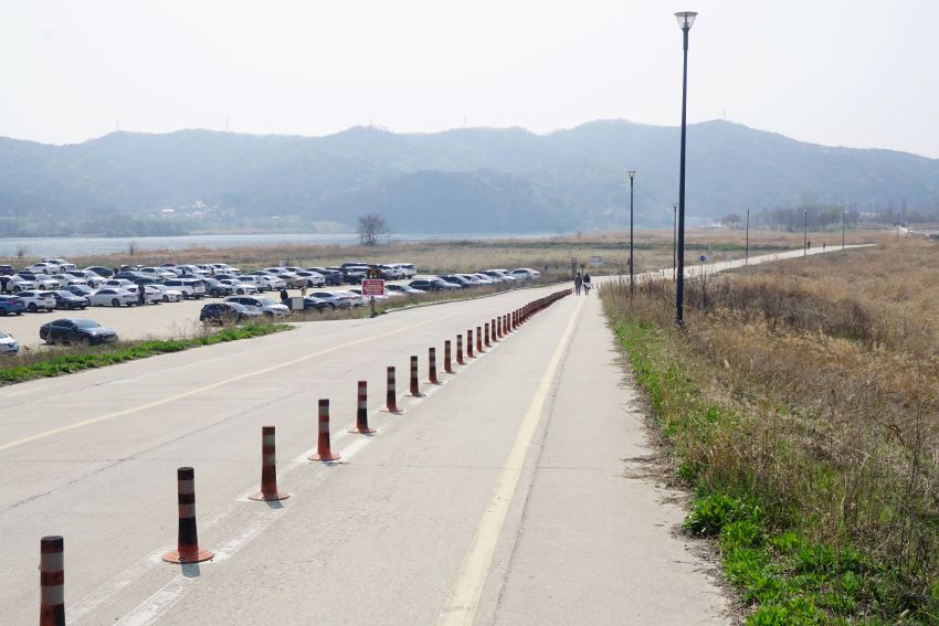 강천섬으로 가는 자전거길, 왼족에 주차장, 거의 만차, 좌 우에 잡초 초원, 자전거길 가운데 중간 분리봉, 뒤로는 야산, 흐린 하늘,