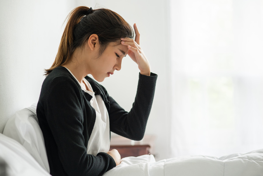 스트레스를 많이 받고 있는 젊은 여성이 침대에 앉아서 왼쪽 손을 머리에 대고 고통스러워 하고 있는 사진