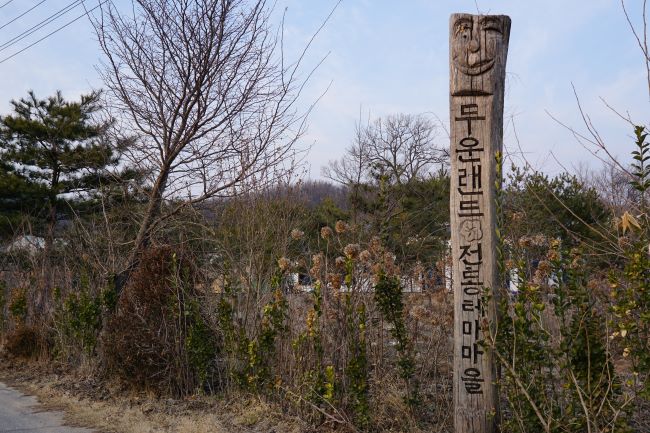 가운데 우뚝 선 장승모양의 나무기둥에 새긴 안내문 검은 글씨, 뒤로는 정원의 잡목,