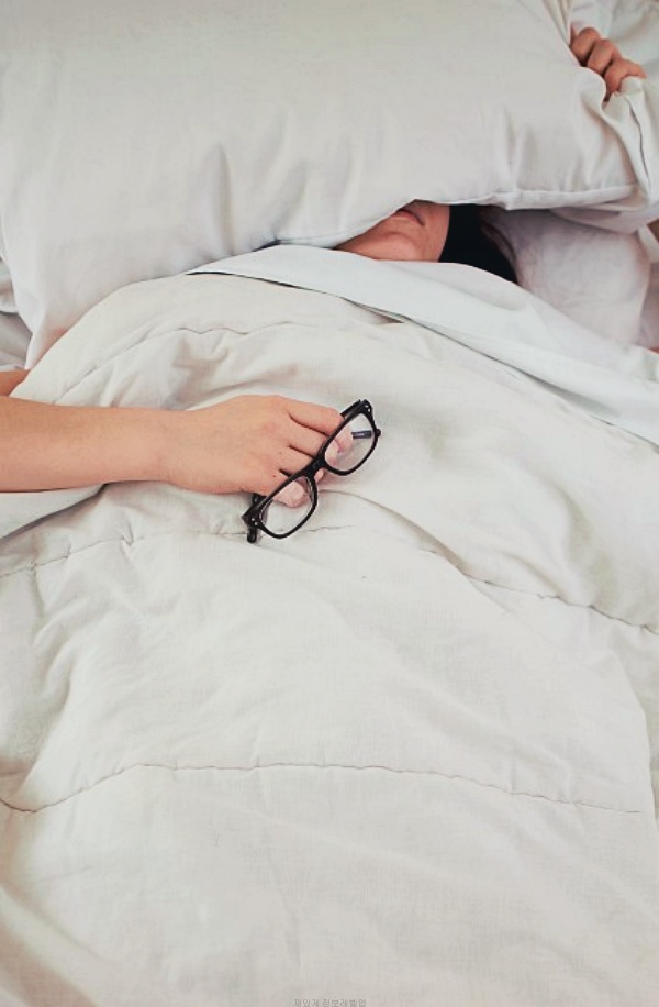 침대위에서 한 손에는 안경을 쥐고 머리위에 베개를 놓아서 피곤함을 표현하고 있는 여자
