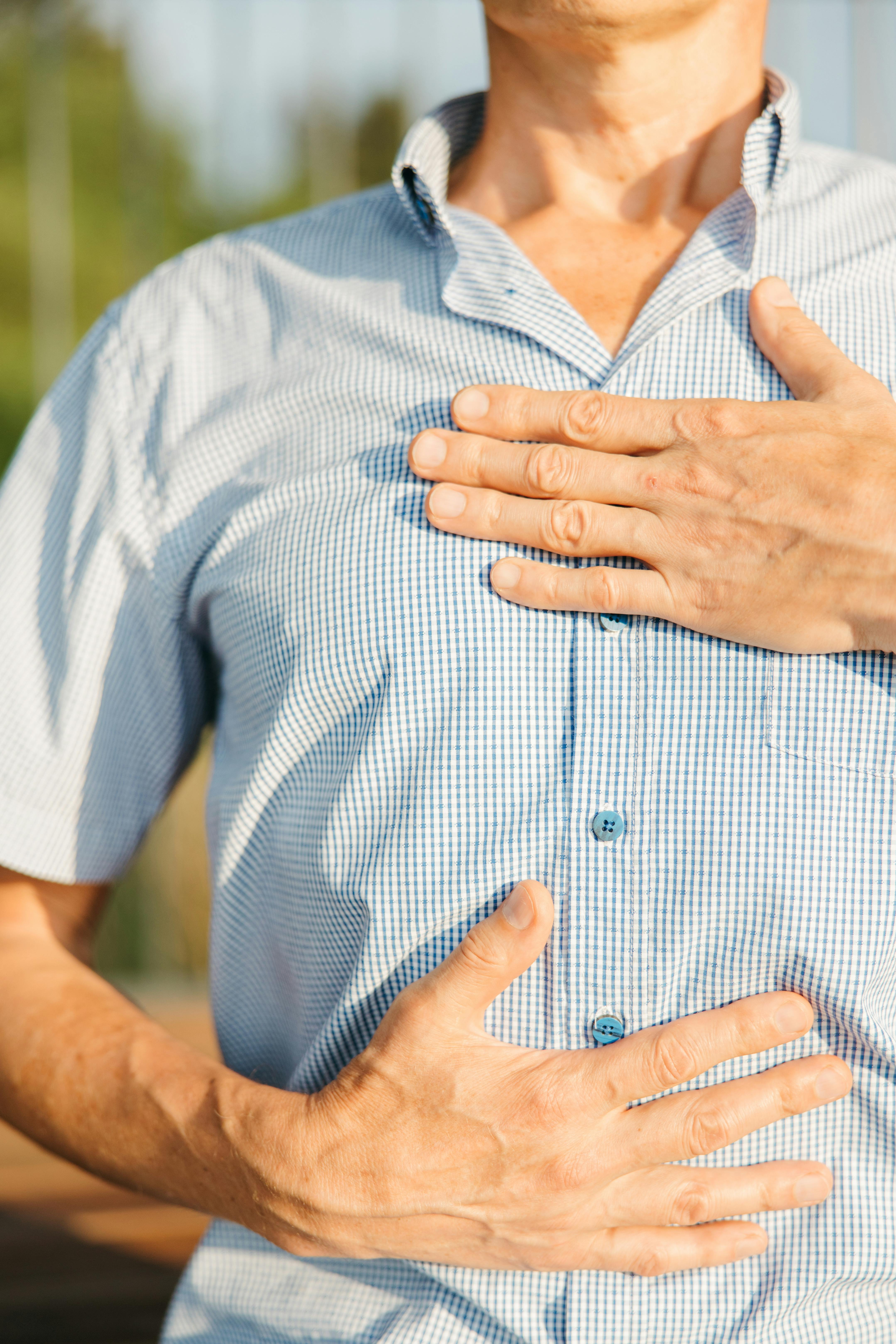 A mid-section shot of a person with their hands gently placed over their chest, wearing a light blue patterned shirt, possibly engaged in a moment of self-soothing or meditation