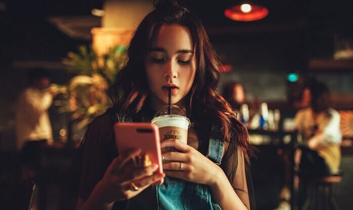 어둑한 카페 안, 한 소녀가 커피를 마시며 다른 한 손으로는 핸드폰을 보고 있는 모습