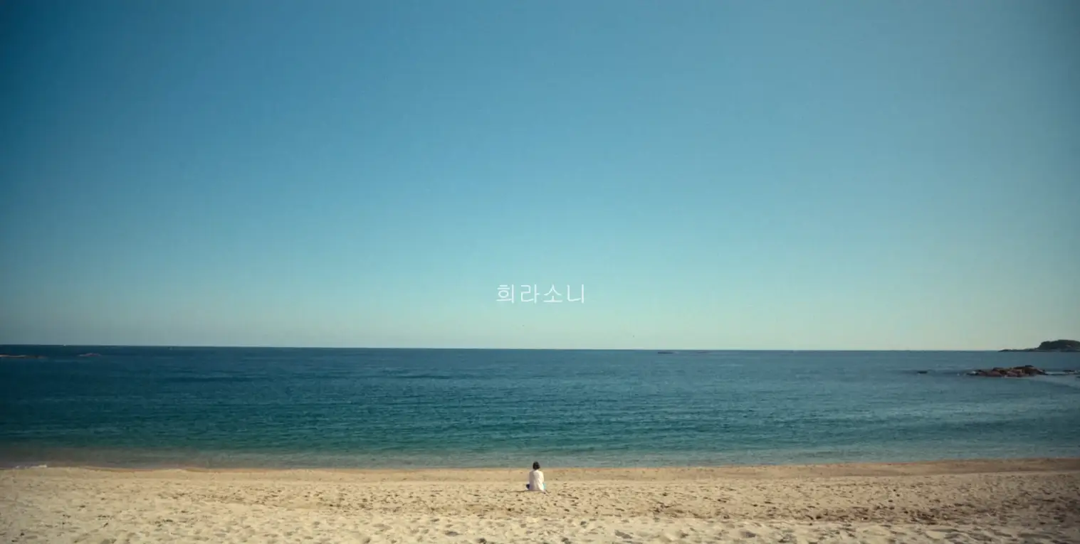 파란 하늘과 파란 바다 앞 모래 사장. 그리고 거기에 조그맣게 앉아 있는 이나영의 뒷 모습.