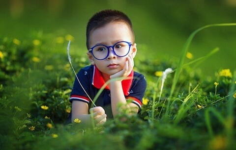 네다섯살 정도 되어 보이는 남자 아이가 파란색 테의 안경을 쓰고 풀밭에 앉아있는 모습