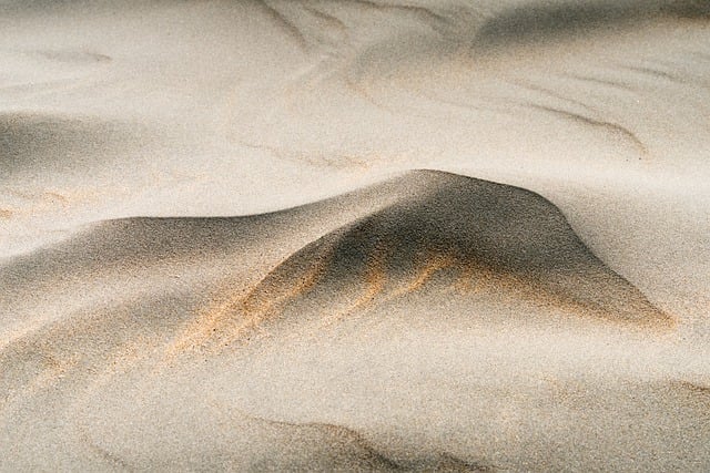 바람에 부드럽게 흩날리는 사막의 모래결을 담은 이미지. 시간과 존재의 덧없음을 상징하는 장면.