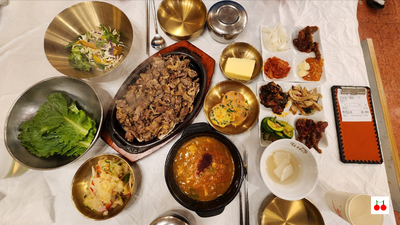 👉 덕소숯불갈비 상차림 전경. 숯불갈비, 반찬들, 찌개, 샐러드, 상추 등이 가지런히 놓여 있음. 고급스러운 놋그릇들이 돋보이는 깔끔한 상차림.