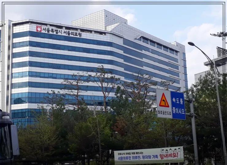 서울특별시 서울의료원 외관 전경. 파란 유리창이 돋보이는 고층 병원 건물 앞에 ‘속도를 줄이시오’라는 도로 표지판과 ‘서울의료원 의료진, 환자와 가족 힘내세요!’라는 응원 현수막이 함께 보임