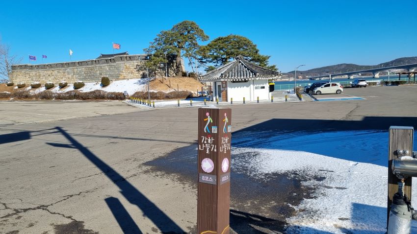 푸른 하늘&#44; 초지진 성곽&#44; 성곽 위에 나부끼는 깃발&#44; 그 앞 큰 소나무&#44; 가운데 팔각 기와지붕의 해설사의 집&#44; 흰 벽&#44; 우측에 주차된 흰색 승용차&#44; 그 우측으로 보이는 초지대교&#44; 강건너 김포&#44;