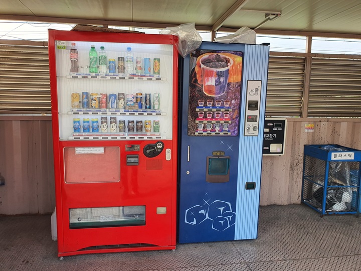 빨간색의-음료수-자판기와-종이컵-사진이-있는-커피-자판기가-나란히-위치해-있는-모습