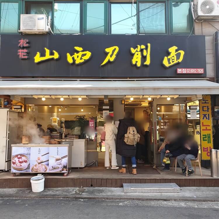 건대입구역에-있는-송화산시도삭면-본점-간판과-입구가-보이고-입구-앞에서-사람들이-웨이팅-하고-있다