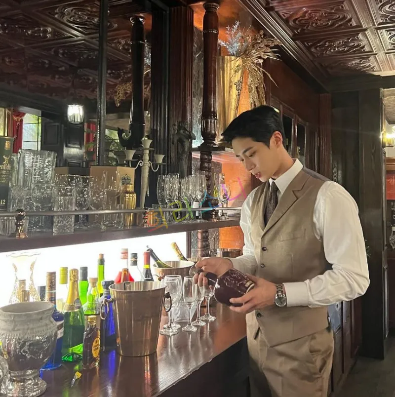 브라운톤의 고급스러운 바(bar)에서 추영우가 베이지색 정장 조끼와 넥타이를 매고 와인병을 들고 있는 모습. 배경에는 다양한 유리잔과 와인병이 장식되어 있다. 추영우는 집중한 표정으로 와인병을 살펴보며 우아한 분위기를 자아낸다.