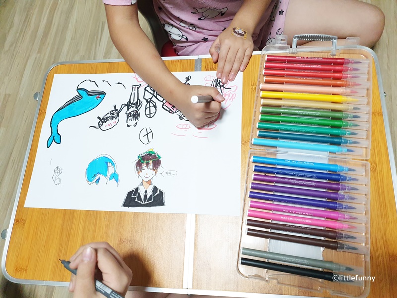 24색 붓펜을 펼쳐놓고 테이블 위 도화지에서 그림을 그리고 있는 아이의 모습