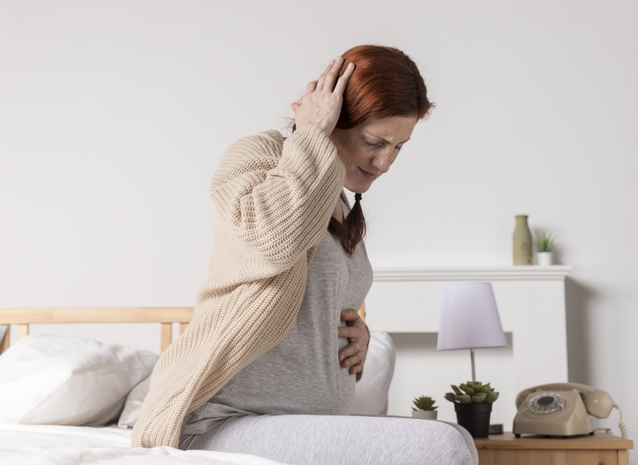 임신을 한 서양 여성이 침대에 앉아서, 뒷목이 뻣뻣해지는 두통으로 인해서 발생되는 통증을 억제하기 위해서 오른쪽 손으로 뒷목을 잡고 있고, 왼쪽 손으로는 배를 대고 있는 사진