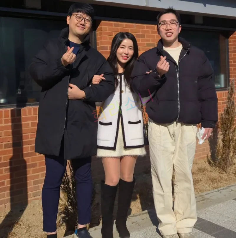 세 명의 사람이 야외에서 사진을 찍고 있다. 가운데 서 있는 여성은 10기 영숙이며 하얀 털이 달린 짧은 코트를 입고 검은색 부츠를 신고 있다. 그녀는 두 남성의 팔을 잡고 서 있으며&amp;#44; 두 남성은 검은색 패딩과 베이지색 바지를 입고 있다. 두 사람 모두 손가락 하트 포즈를 취하고 있다. 배경은 붉은 벽돌 건물과 창문이 보인다. 자막에는 &amp;quot;희라소니&amp;quot;라는 텍스트가 보인다.
