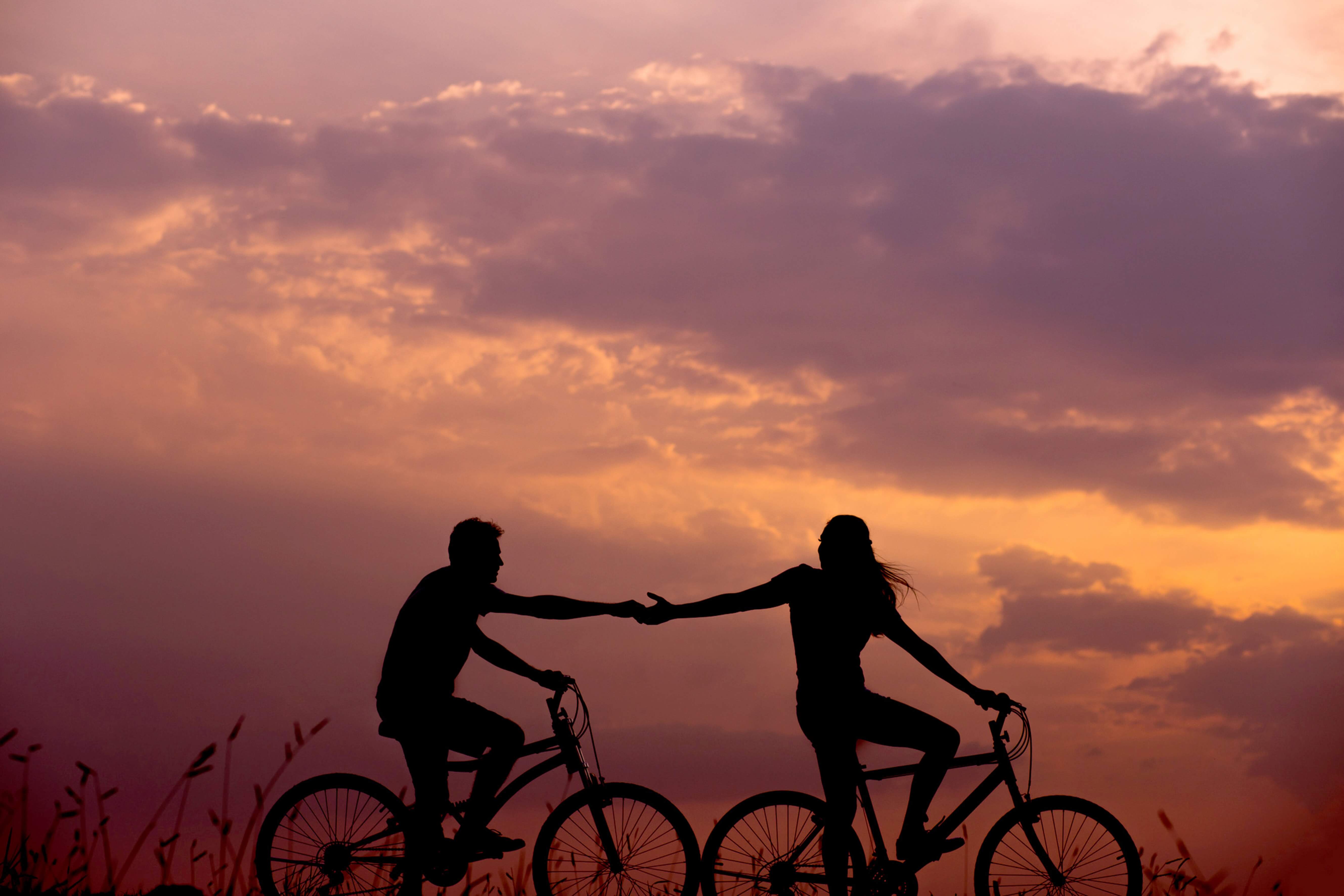 노을진 하늘 앞에서 여자와 남자가 자전거를 타고 서로의 손을 잡고있는 그림자