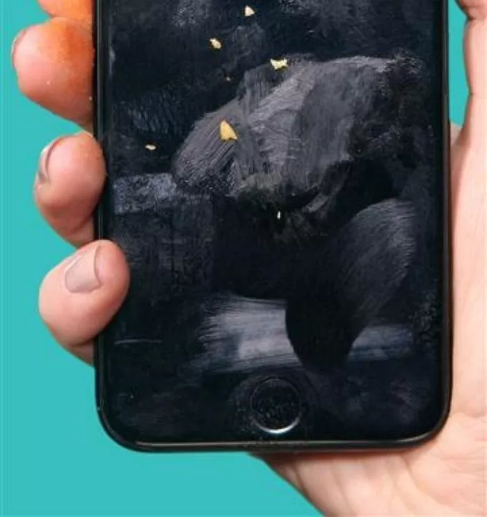 지문과 유분으로 덮인 스마트폰 화면의 모습. 검은색 스마트폰을 손에 들고 있으며, 화면 전체에 지문 자국이 번들거리고 여러 개의 얼룩과 자국이 선명하게 보인다. 화면 상단에는 작은 먼지나 이물질도 묻어 있다. 배경은 청록색이다.
