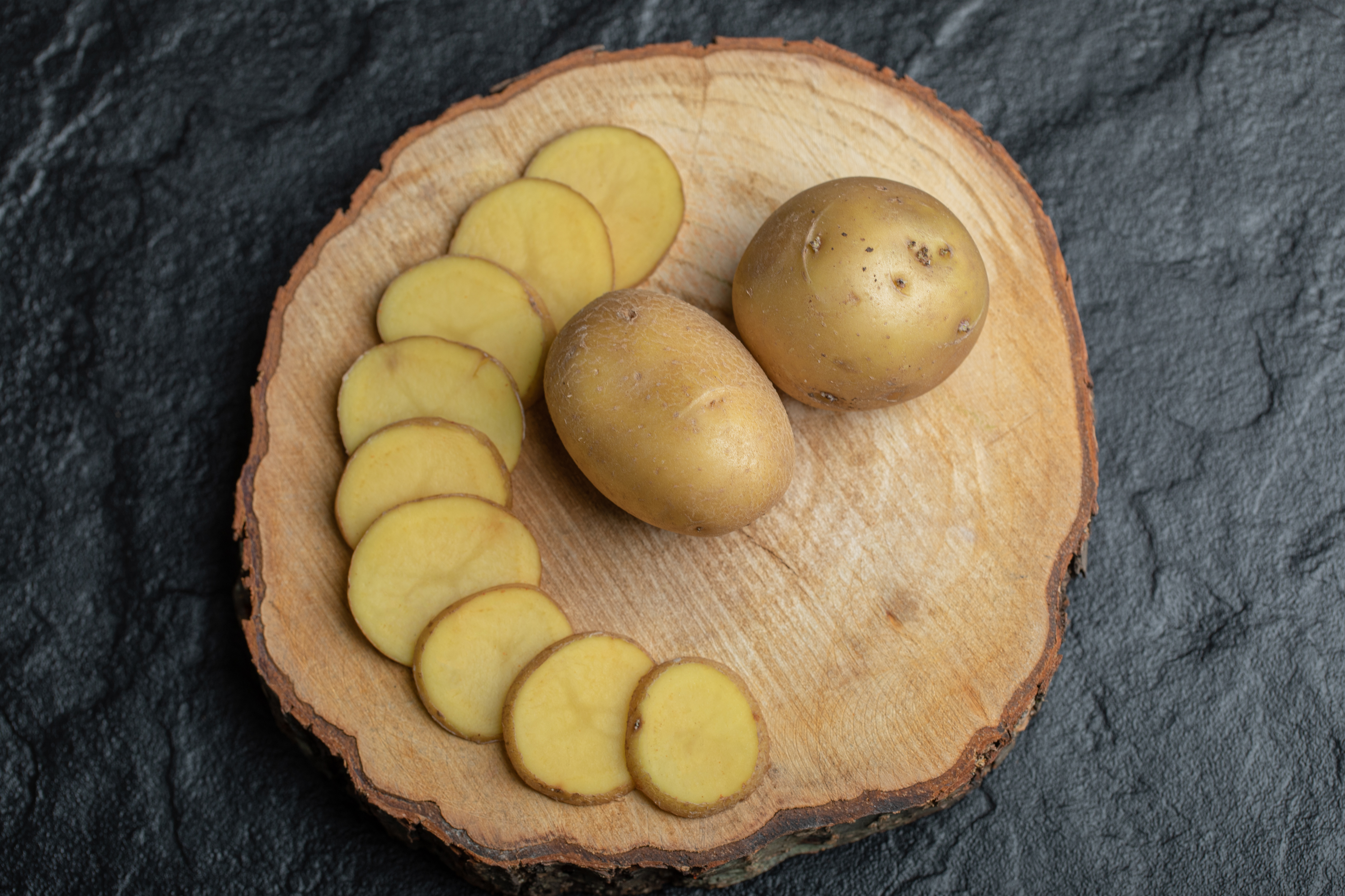 야생에서 채취를 한 감자를 검은 바닥 위에 동그란 나무 깔판 위에 통으로 2개를 놓고 그 옆으로 감자를 편으로 썰어놓은 것을 차례대로 포개서 놓아 두고 위에서 찍은 사진 (sliced-whole-potato-brown-wooden-board)