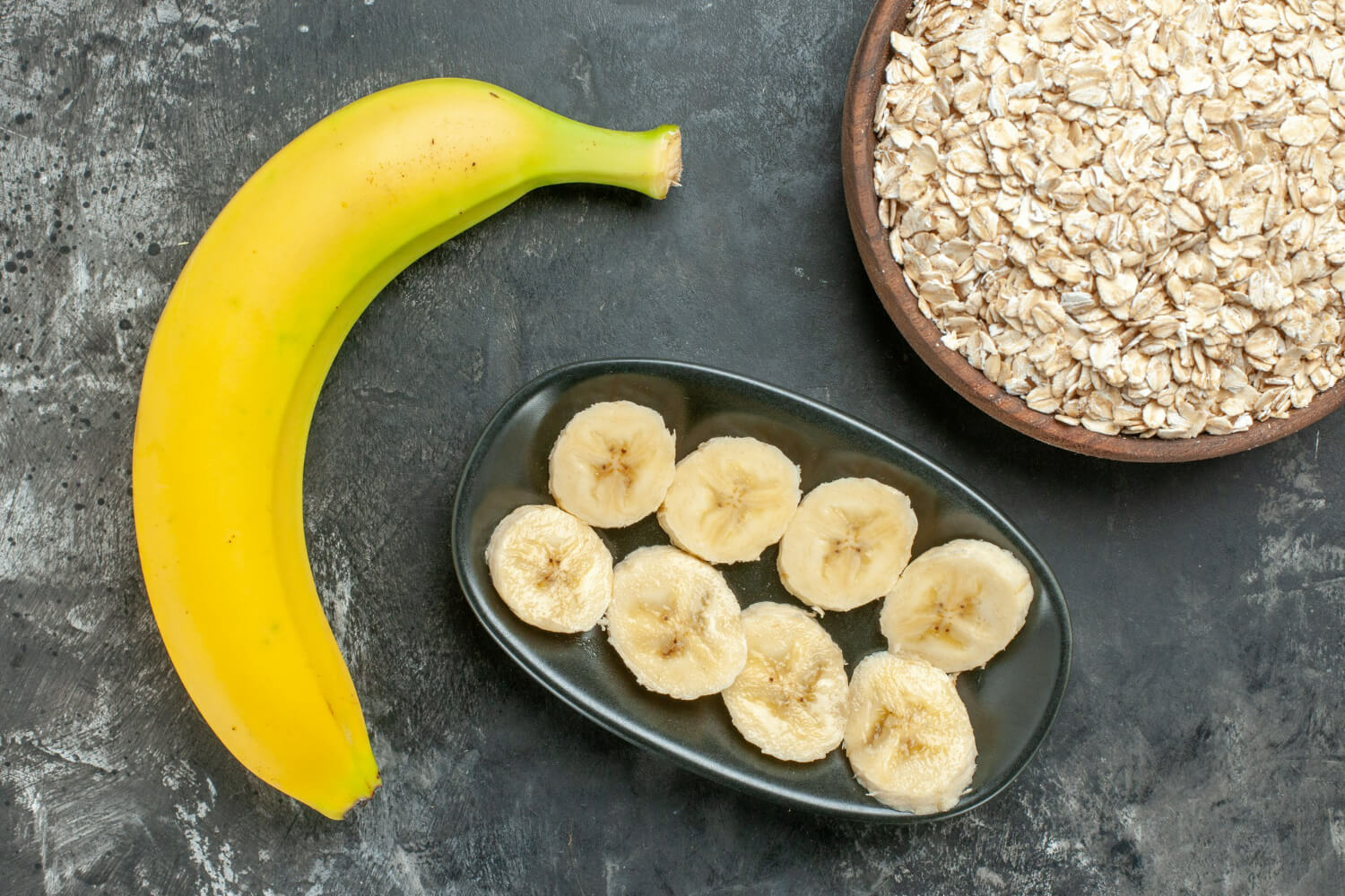 바나나 한개와 검은 접시에 잘라진 바나나가 놓여져 있고 그 옆에 귀리가 담겨있는 접시가 놓여져 있다.