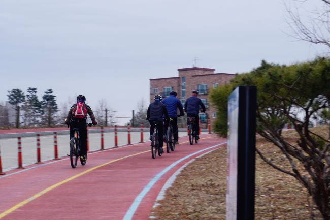 고구마색 자전거길을 달리는 자전거 4&#44; 모두 남성&#44; 뒷모습&#44; 건너편에 붉은 벽돌 건물&#44; 푸른 하늘&#44;