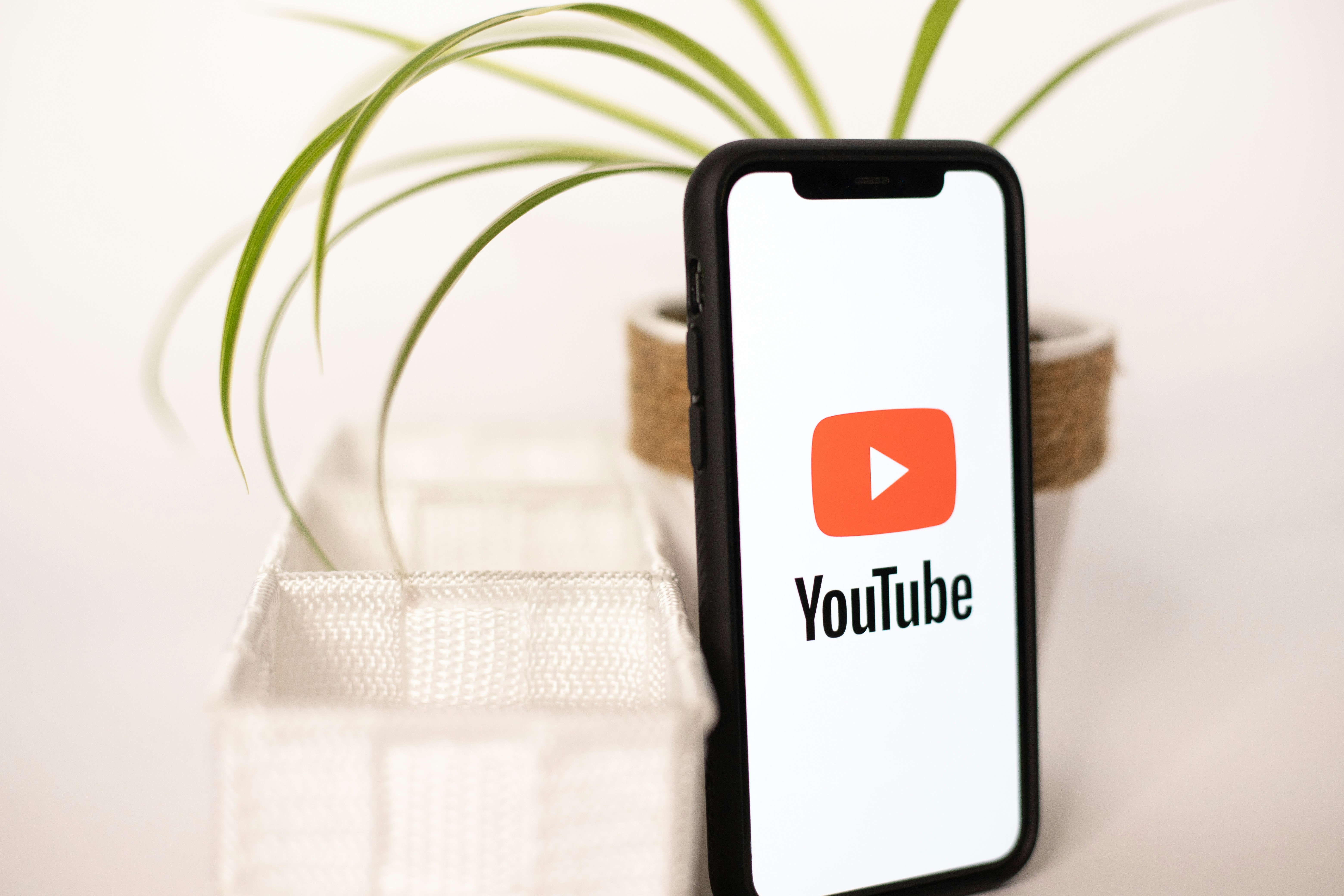 A smartphone displaying the YouTube logo, placed on a white table with a plant in the background. Ideal for content about YouTube Shorts, mobile video creation, and digital monetization.