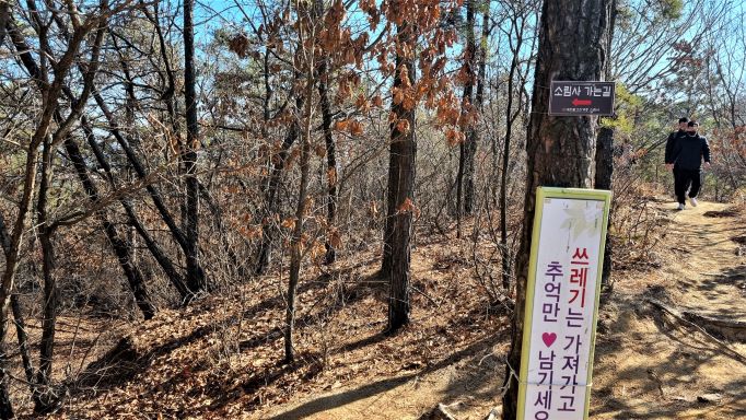 우거진 참나무 숲&#44; 삼거리&#44; 나무줄기에 붙은 소림사 가는길안내 화살표&#44; 쓰레기 버리지 말고 추억만 남기세요 라는 안내문&#44; 우측 산길에서 남성 2 걸어오고 있음&#44;