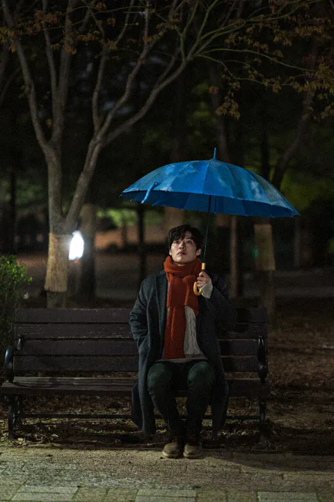 영화 비와 당신의 이야기의 강하늘이 파란 우산을 쓰고 붉은 목도리를 두른채 공원 벤치에 앉아있다.