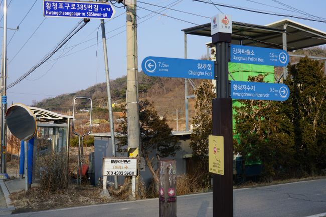 창후항 가는 이정표, 푸른 바탕에 흰글씨, 그 아래에 나들길 이정표 말뚝, 가운데에 회색 전봇대, 좌측에 버스정류장, 둥그런 반사경, 뒤로 보이는 야산,