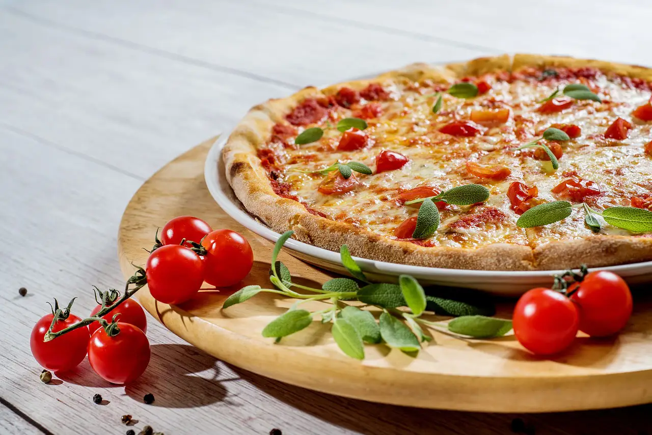 쿠팡- 흰 나무테이블 위 빨간 방울토마토 여러개가 놓인 나무쟁반 위 흰접시 안 붉고 초록색 잎이 덮여있는 피자
