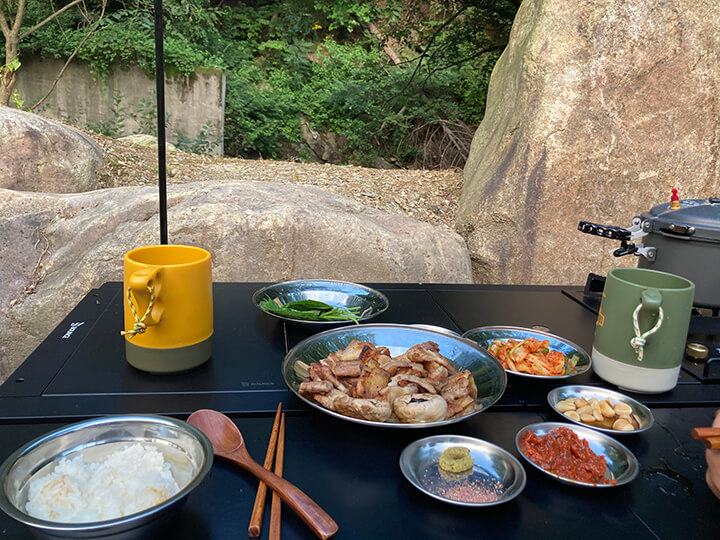 계곡과-숲을-배경으로-캠핑테이블-위-스텐접시에-담겨진-음식들