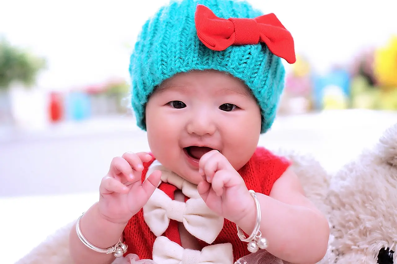 부모급여- 빨간 리본이 달린 파란모자를 쓰고 왼손가락은 입에 넣고 웃는 아기