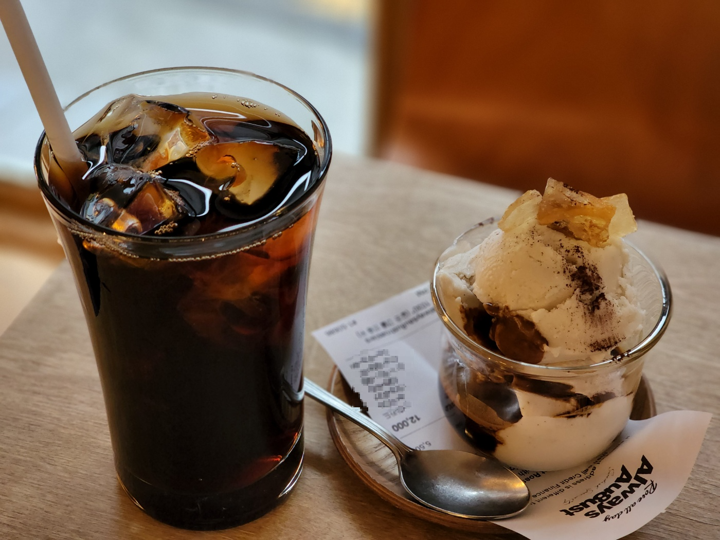 긴 투명한 컵에 가득담긴 아이스커피 한잔과 그 옆에 나무 코스터와 작은 투명한 잔 사이에 영수증을 끼워 놓은 작은 스푼과 함께 제공된 코르타도 아이스크림 메뉴