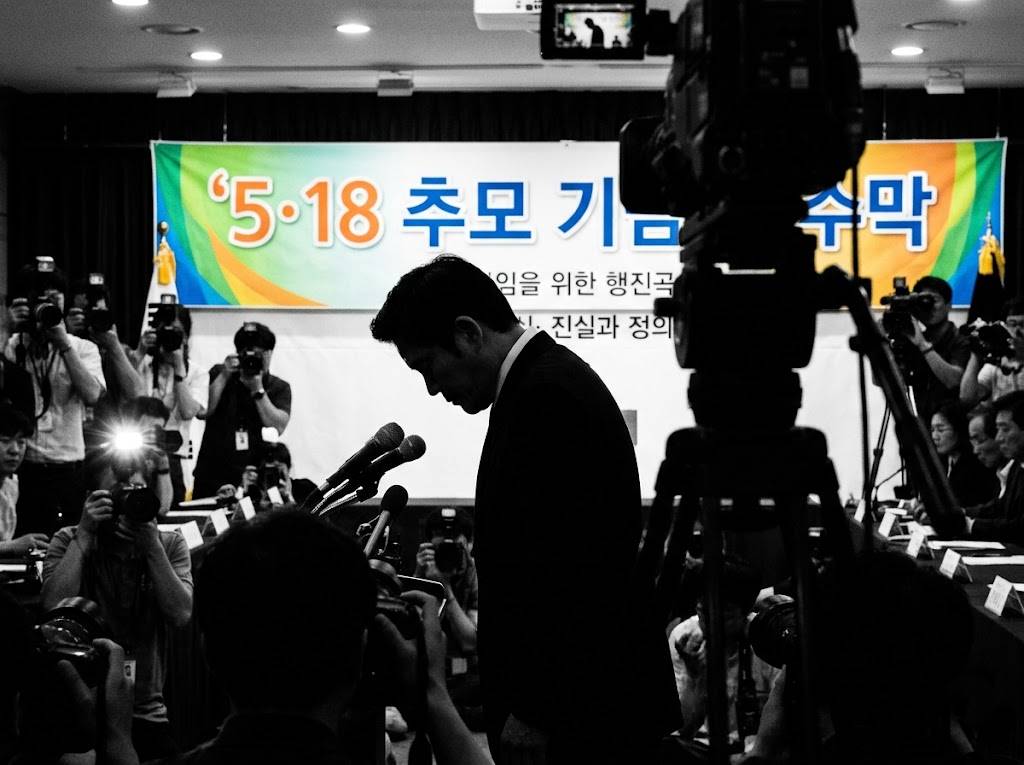 기자회견장에서 고개를 숙인 기업 임원 실루엣과 배경에 5&middot;18 추모 기념 현수막이 걸린 장면, 흑백과 컬러 혼합 구도