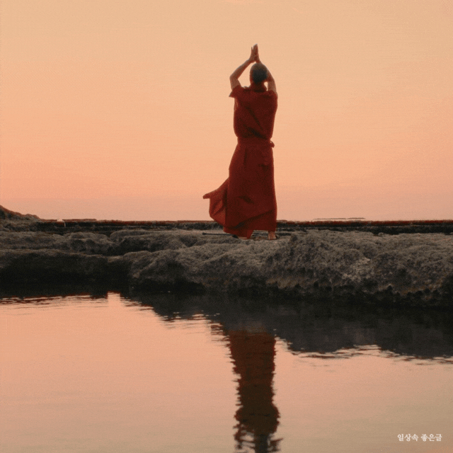 분홍빛 노을 아래 붉은 옷을 입은 사람이 기도하듯 두 손을 들고 서 있는 고요한 장면의 실루엣