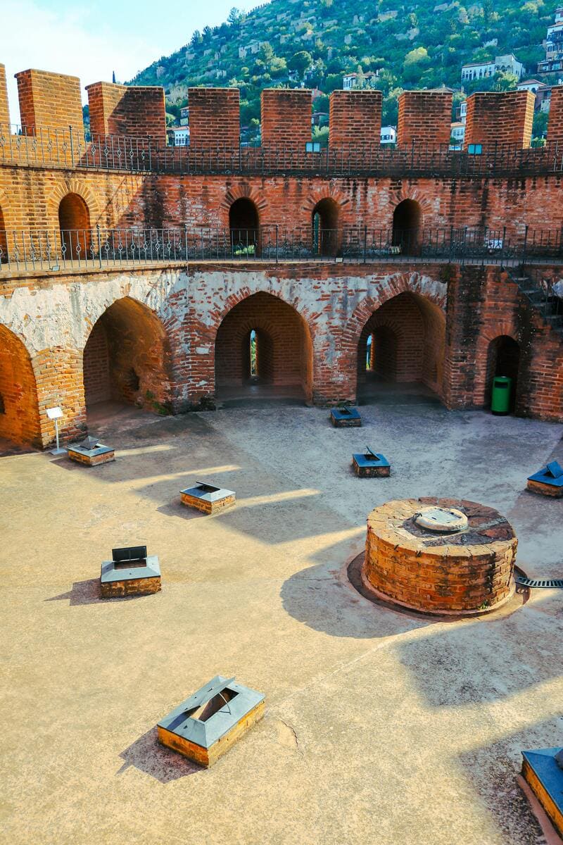 알라라 성채 내부의 원형 구조. 바닥에는 벽난로로 추정되는 구조물과 벽을 따라 설치된 벽감이 보이며, 위층에는 아치형 출입구와 방어용 틈새가 정렬되어 있습니다. 뒤로는 푸른 산과 마을이 내려다보입니다.