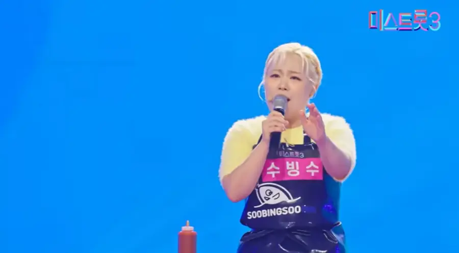 미스트롯3- 파란색배경 노란색 웃옷 파란앞치마를 입고 가슴에 분홍바탕 흰글씨 수빙수 이름을 달고 마이크를 잡고 앞을 보며 노래하는 모습