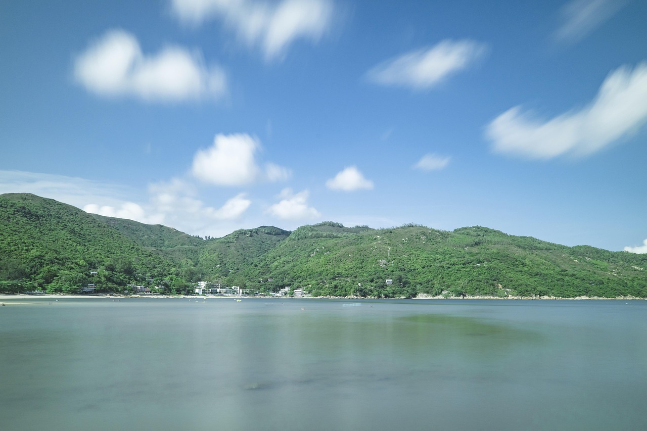 짙은 녹음을 두른 산자락이 부드럽게 이어지고, 그 아래로 잔잔하게 흐르는 호수 같은 바다가 넓게 펼쳐져 있다. 하늘엔 흰 구름들이 천천히 이동하며 고요한 시간의 흐름을 암시하고, 맑고 푸른 하늘은 자연의 여유로움을 더한다. 멀리 작은 건물들이 드문드문 자리해 자연 속에 녹아든 인간의 흔적을 보여주며, 전반적으로 청명한 공기와 평화로운 분위기가 감도는 한적한 해안 마을의 풍경을 담은 사진.