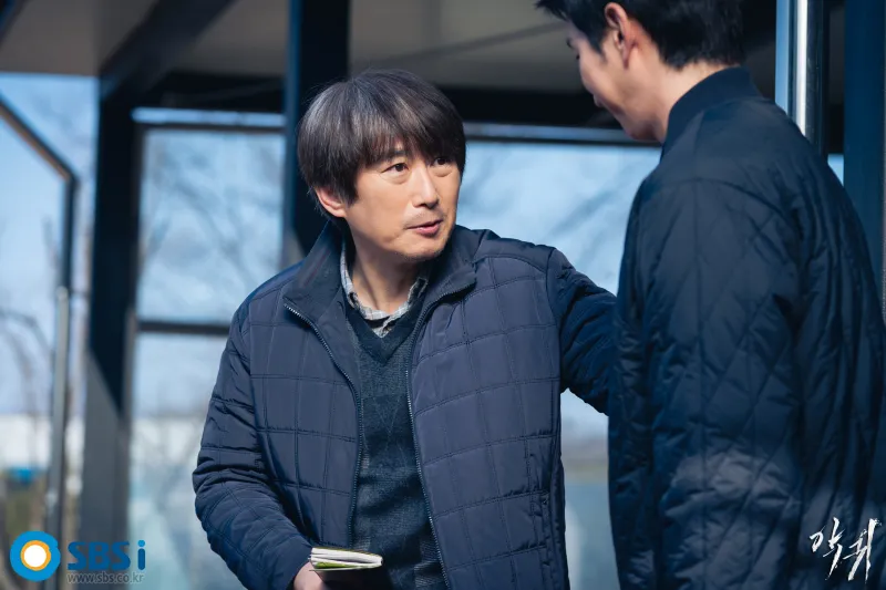 한 시골 건물에서 수사 수첩을 들고 누군가와 심각하게 이야기하고 있는 서문춘