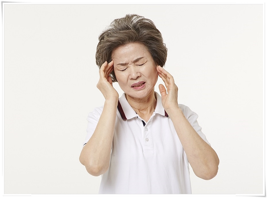 양쪽 관자놀이 부근에 손을 가져다 단 채 두 눈을 감고 찡그린 표정을 짓고 있는 고령 여성의 모습