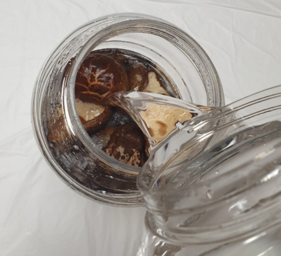 하얀 바닥 위에 투명한 유리병에 표고버섯을 반쯤 담아 두고 청주를 그 속에 부어 표고버섯 술을 만들고 있는 사진