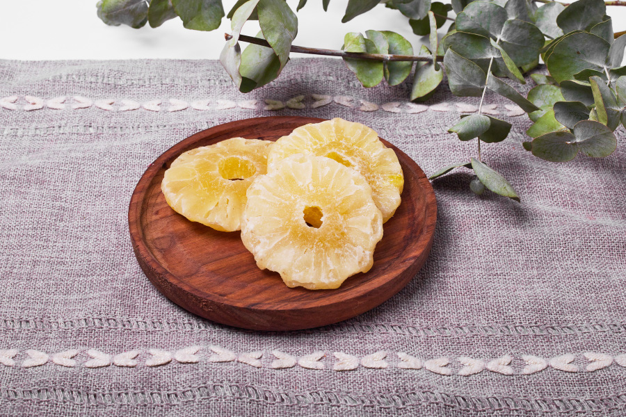 야생에서 채취를 한 파인애플을 편으로 얇게 썰은 것을 바싹 말린 것을 테이블 위에 고동색의 동그란 나무 접시에 3개 놓아두고, 뒤에는 나뭇가지를 놓아두고 찍은 사진