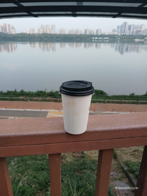 아침 한강 전망을 배경으로 난간 위에 놓인 테이크아웃 커피 컵