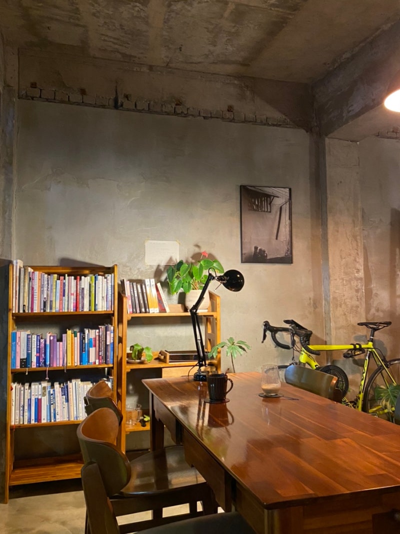 콘크리트 벽의 조용한 공간, 책장이 놓인 방 안에 긴 나무 테이블과 스탠드 조명이 켜진 쉼표의 서재 풍경