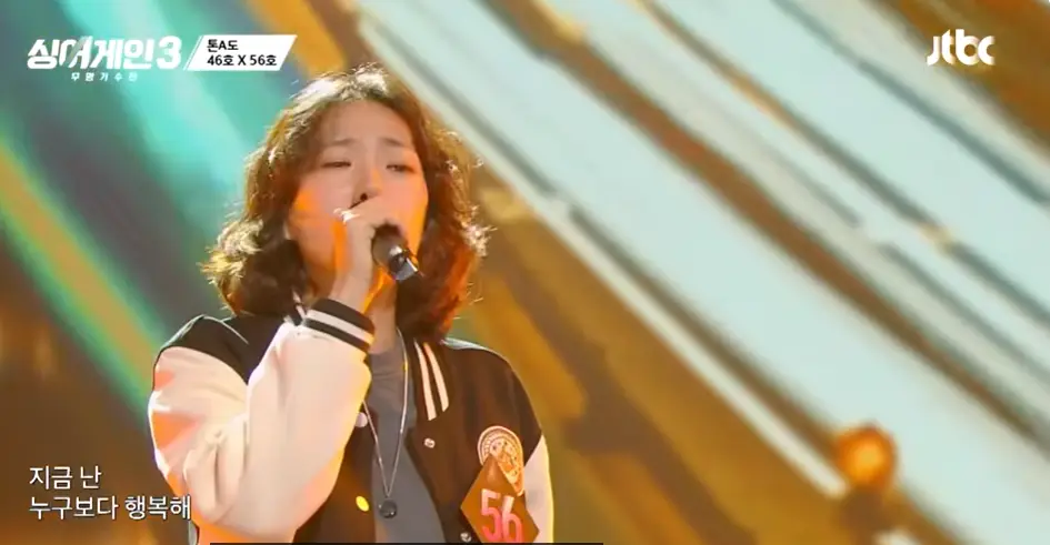 싱어게인 3- 검정색 몸통에 흰색팔부분 점퍼를 입고 단발 곱슬머리상태로 오른손으로 마이크를 쥐고 노래하는 56호 상반신