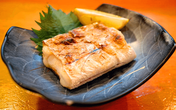 흑회색 접시에 갈치구이 한 조각이 있다. 접시는 위쪽으로 살짝 구부러졌고, 갈치구이 위에는 레몬 한조각이 있다.