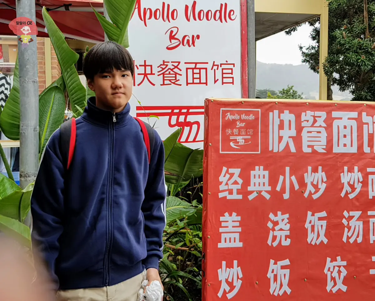 호주-멜번-그레이트-오션-로드-아폴로-베이-몰-아폴로-누들-바-아시아요리-음식점-간판-앞에서-포즈를-취하고-있는-소년